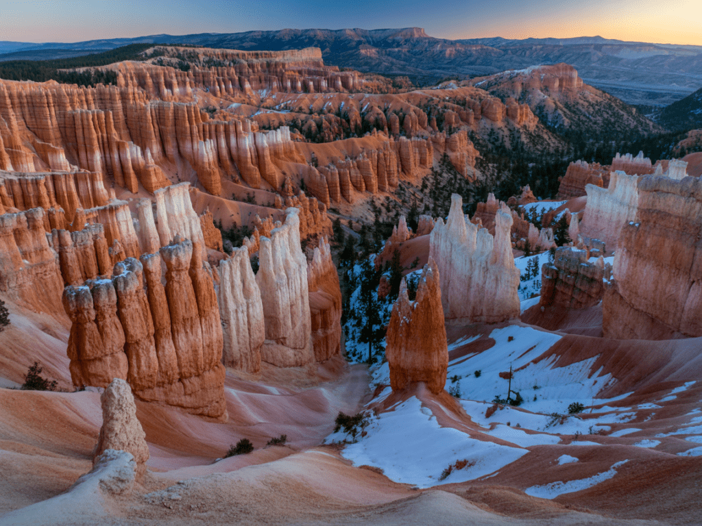Bryce Amphitheater from Inspiration Point at sunrise, thousands of orange and red hoodoo spires rising from amphitheater floor dusted with snow, warm golden first light raking across the formations from the east, dramatic contrast between lit spires and deep shadow, Utah high plateau landscape photography